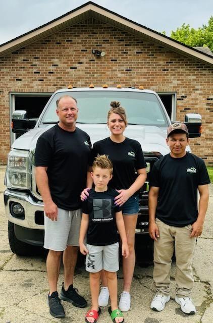 Group of people standing in front of a large pickup truck and a brick garage.